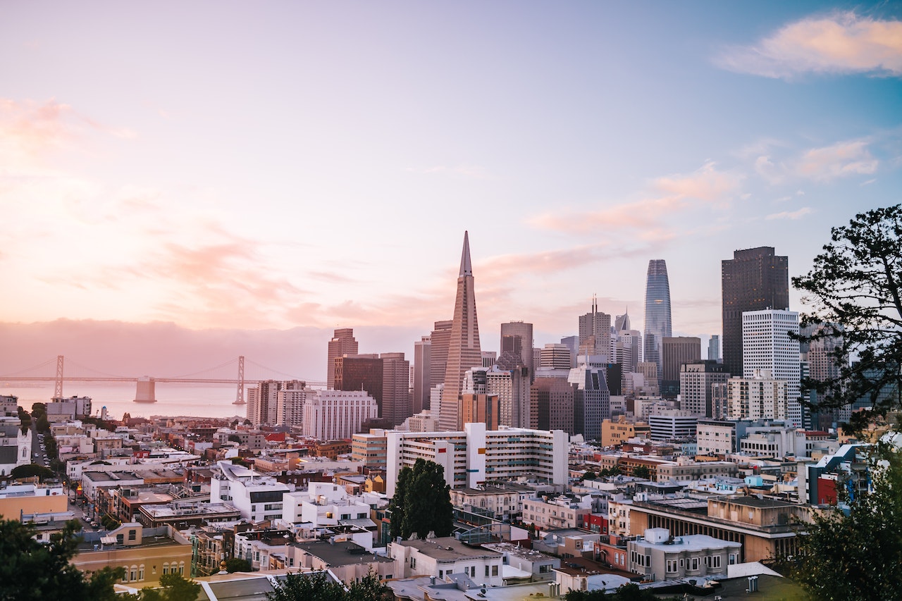 A view of downtown San Francisco, with the Transamerica Pyramid and Salesforce Tower visible, and the Bay Bridge in the background. The sky is awash in gentle golden light, with a boundary of fog seen past the Bay Bridge.